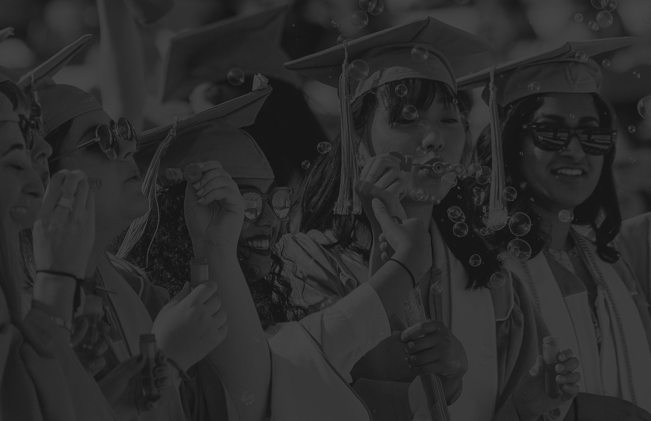 Photo of female graduates at commencement blowing bubbles with black overlay on top of the photo.